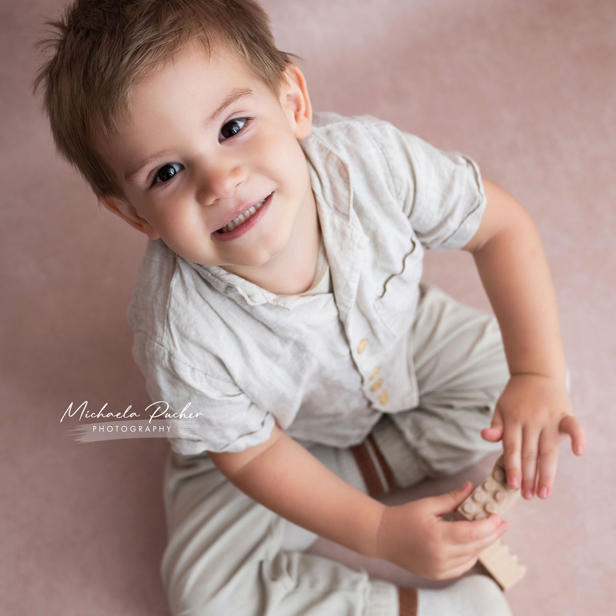 Lachender kleiner Junge sitzt am Boden auf einem rosigen Hintergrund und spielt mit einem Holzbaustein, während er direkt in die Kamera blickt.